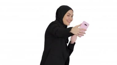 Beyaz arka planda cep telefonuyla fotoğraf çeken tesettürlü gülümseyen Arap kadın..