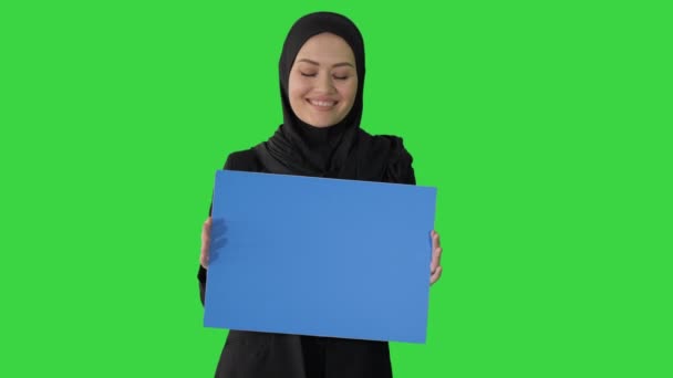 Улыбающаяся арабка в хиджабе держит чистый голубой плакат и смотрит в камеру на зеленом экране, Chroma Key. — стоковое видео