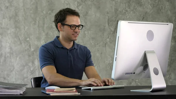 Giovane uomo sorridente seduto alla sua scrivania in ufficio e al lavoro — Foto Stock