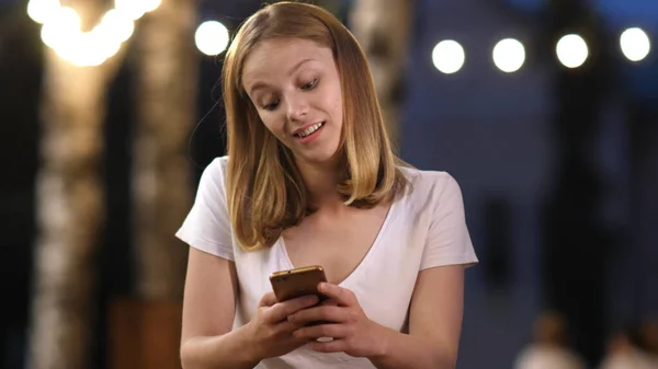 Giovane attraente ragazza interessata sta digitando il messaggio sul suo smartphone in serata in estate, sorridente, concetto di comunicazione. — Foto Stock