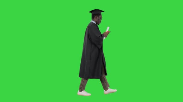 Afričtí američtí studenti v promoční župan textování na telefonu při chůzi se svým diplomem na zelené obrazovce, Chroma Key.