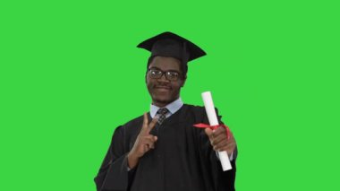 Mezuniyet cüppeli mutlu Afro-Amerikan erkek öğrenci diploması ile Yeşil Ekran, Chroma Key 'de kamera önünde poz veriyor..
