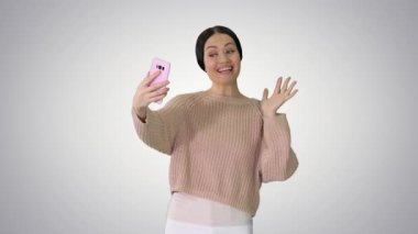 Gülümseyen genç bir kadın telefonunda video görüşmesi yapıyor gradyan arka planda el sallıyor..