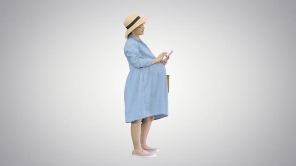 Joyeux femme enceinte en robe bleue et chapeau en utilisant son smartphone sur fond dégradé. — Video