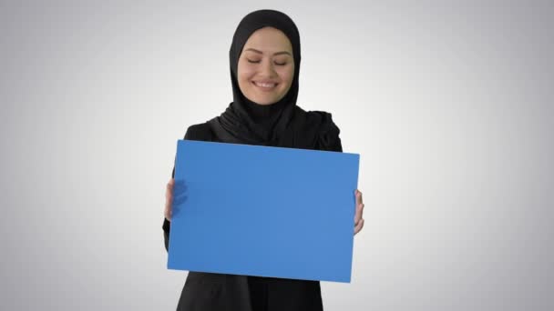 Улыбающаяся арабка в хиджабе держит чистый голубой плакат и смотрит в камеру на градиентном фоне. — стоковое видео