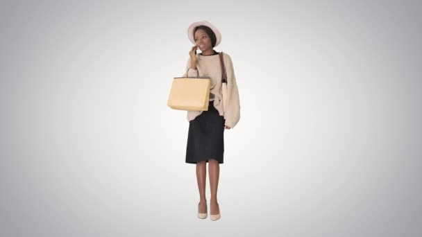 African american girl in oversized knitwear and hat talking on her phone while walking with shopping bags on gradient background. — Stock Video