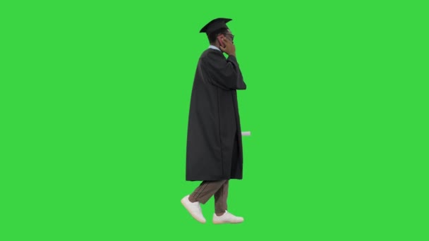 Улыбающийся африканский студент-американец в выпускном халате разговаривает по телефону во время прогулки по зеленому экрану, хрома-ключ. — стоковое видео