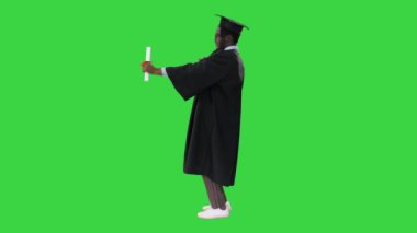 Mezuniyet cüppeli mutlu Afro-Amerikan erkek öğrenci diploması ile Yeşil Ekran, Chroma Key 'de kamera önünde poz veriyor..