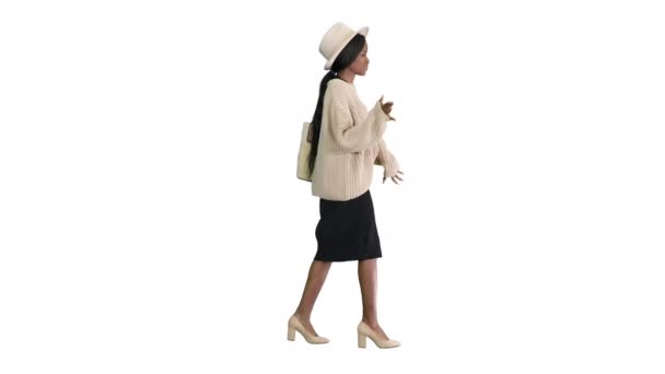 Elegante mujer afroamericana en punto y sombrero blanco bailando mientras camina sobre fondo blanco. — Vídeo de stock