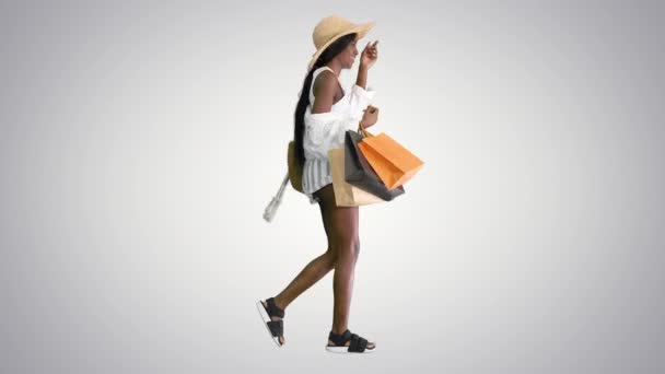 Счастливая африканская американка в соломенной шляпе ходит с пакетами для покупок на градиентном фоне. — стоковое видео