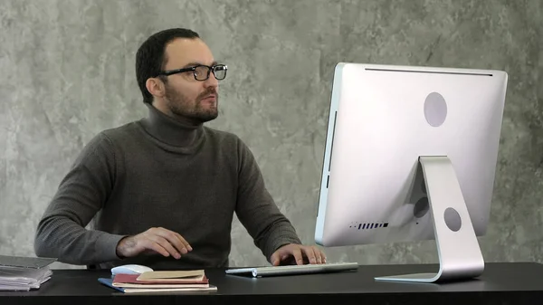 Злой человек сидит за столом с компьютером над серой backgroun — стоковое фото