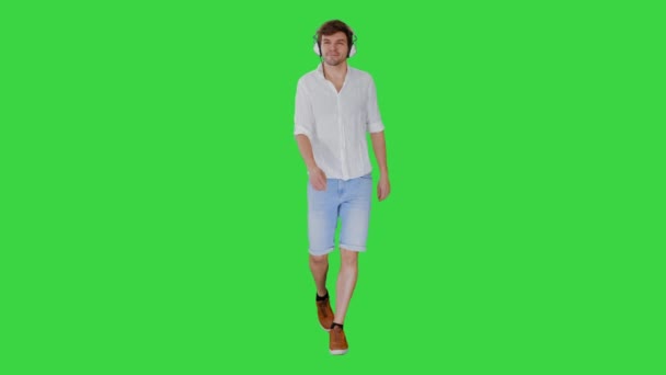 Lässiger junger Mann, der Musik über Kopfhörer hört, während er geht und auf einem Green Screen in die Kamera schaut, Chroma Key. — Stockvideo