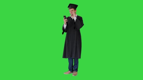 Ridendo studente maschio in abito di laurea e mortai avendo videochiamata sul suo telefono su uno schermo verde, Chroma Key. — Video Stock