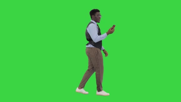 緑の画面、クロマキーを歩いている間に彼の手に携帯電話を保持しているビデオ通話を行うアフリカ系アメリカ人ビジネスマンの笑顔. — ストック動画