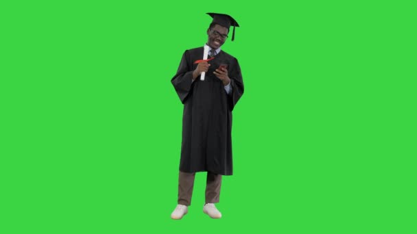 Χαμογελώντας αφροαμερικανός φοιτητής με στολή αποφοίτησης με δίπλωμα γραπτών μηνυμάτων στο τηλέφωνο σε μια πράσινη οθόνη, Chroma Key. — Αρχείο Βίντεο