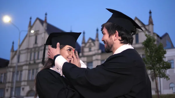 Twee gelukkige afgestudeerden. Mannelijke afgestudeerde student helpt het meisje om de pet recht te zetten. Ze lacht naar hem.. — Stockfoto