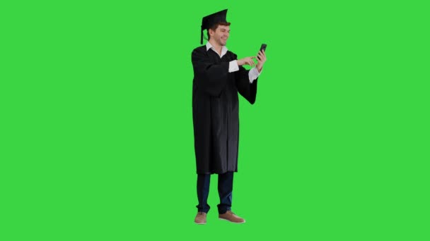 Glückliche männliche Absolvent in Kleid und Mörtel mit emotionalen Videoanruf auf seinem Telefon auf einem Green Screen, Chroma Key. — Stockvideo