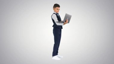 Papyonlu ve yelekli konsantre bir çocuk gradyan arkaplanda dururken dizüstü bilgisayar kullanıyor..