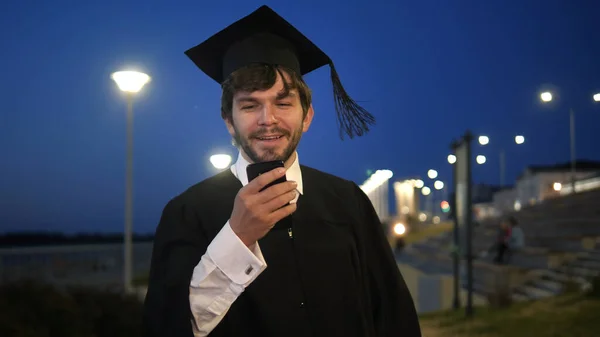 Młody uśmiechnięty mężczyzna absolwent gospodarstwa smartfon smsowanie coś i chodzenie. — Zdjęcie stockowe