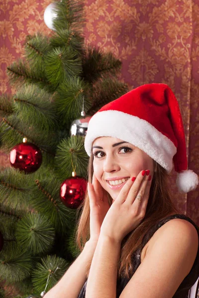 在圣诞树的背景上微笑的女孩在圣诞节帽子微笑 — 图库照片