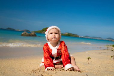 bebek beyaz kum üzerinde mavi deniz kıyısında Noel Baba gibi giyinmiş