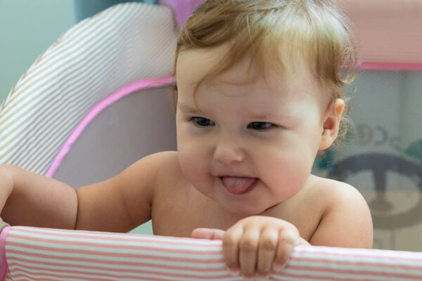 Милый смеющийся ребенок, стоящий в розовой кровати. Розовый питомник для маленьких детей. Маленькая девочка учится стоять в своей кроватке. Ребенок стоит в кроватке в солнечной спальне. Ребенок высунул язык
