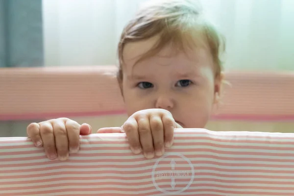 Bonito bebê rindo de pé em uma cama rosa. Viveiro rosa para crianças pequenas. Uma menina aprende a ficar no berço. Um bebê está de pé em um berço em um quarto ensolarado. A criança está triste — Fotografia de Stock