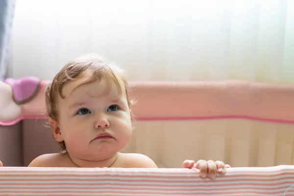 Cute laughing baby standing in a pink bed. Pink nursery for small children. A little girl learns to stand in her crib. A baby is standing in a crib at a sunny bedroom. TThe child is sad and smiling, — Stock Photo, Image