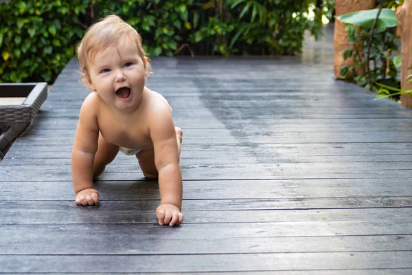 Pretty crawling baby girl smiling and crawling on the wooden brown walkway on the background of green bushes and trees. The child found on the floor and trying to eat. There is a place for text.