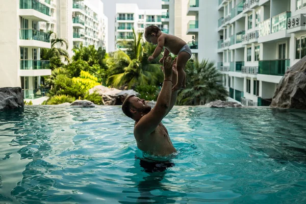 Vader gooit de baby in het zwembad op de achtergrond van kokospalmen. Gelukkig actieve familie jonge vader en zijn schattige dochter schattig peuter meisje spelen in een zwembad springen in het water genieten van — Stockfoto