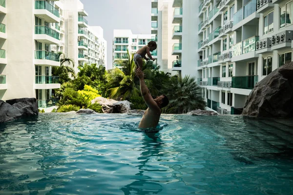 Vader gooit de baby in het zwembad op de achtergrond van kokospalmen. Gelukkig actieve familie jonge vader en zijn schattige dochter schattig peuter meisje spelen in een zwembad springen in het water genieten van — Stockfoto