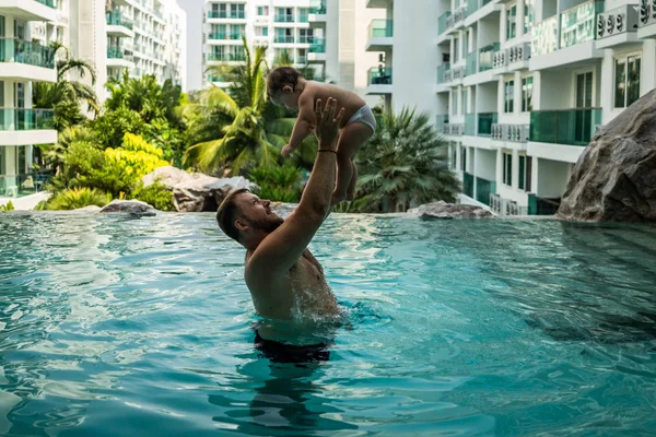 Vader gooit de baby in het zwembad op de achtergrond van kokospalmen. Gelukkig actieve familie jonge vader en zijn schattige dochter schattig peuter meisje spelen in een zwembad springen in het water genieten van — Stockfoto