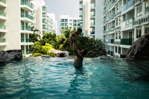 Vader gooit de baby in het zwembad op de achtergrond van kokospalmen. Gelukkig actieve familie jonge vader en zijn schattige dochter schattig peuter meisje spelen in een zwembad springen in het water genieten van — Stockfoto