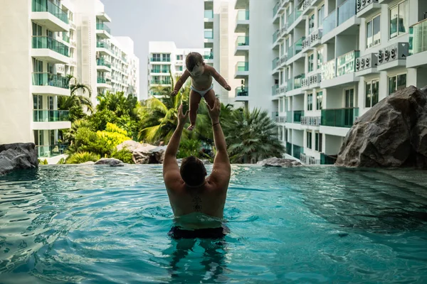 Tata zgłasza dziecko w puli na tle palmy kokosowe. Happy active rodziny młody ojciec i jego córka dziewczynka śliczny maluch gry w basenie, skoki do wody, ciesząc się — Zdjęcie stockowe