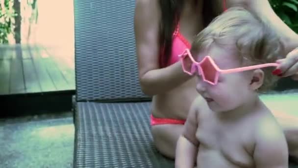 Mamá viste a su hija con gafas de sol, el bebé no quiere usarlas y se quita — Vídeos de Stock