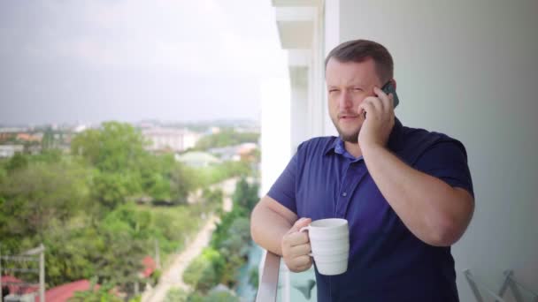 Uomo sul balcone che beve con una tazza e parla al telefono . — Video Stock