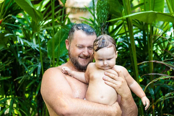 Papà e bambino sotto una doccia tropicale. papà e bambino giocare sulla spiaggia sotto doccia spruzzi d'acqua — Foto Stock