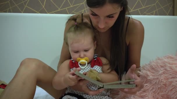 Madre y su linda hija están sentadas en un sofá en la sala de estar. Ellos están leyendo el libro de los niños . — Vídeo de stock