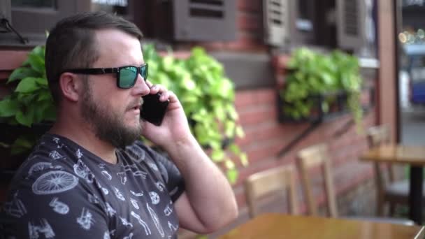 Bebaarde man praten aan de telefoon en chatten in een café op de straat. 4k. — Stockvideo