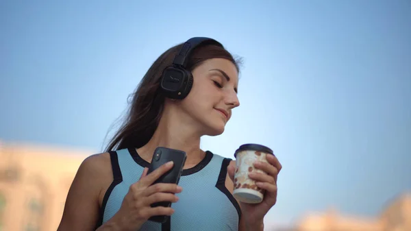 Mooie jonge vrouw met behulp van slimme telefoon, typen van berichten, het luisteren naar muziek, drinken koffie tijdens het wandelen op de stedelijke stad straat. Close-up — Stockfoto