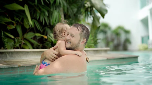 Cute little kid and his father having a swimming lesson in the pool. Father holds daughter in his arms and hugs.