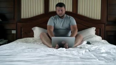 Yatağın üstüne oturan ve dizüstü bilgisayarda çalışan genç erkek. 4k.