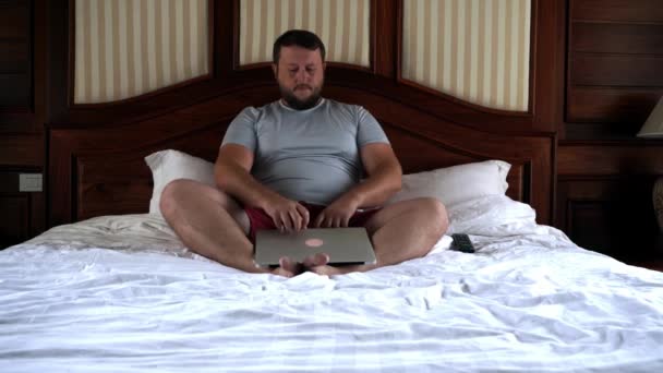 Jonge man zittend op bed en die op laptop werkt. 4k. — Stockvideo