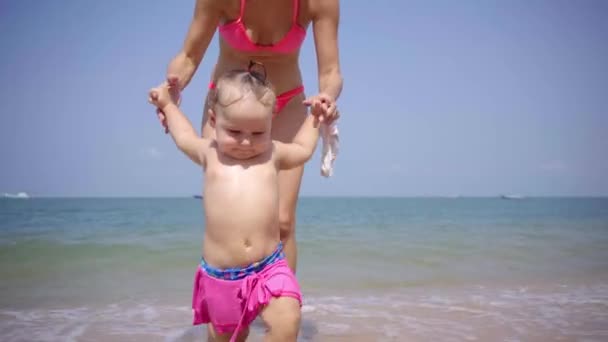 Mamá e hija caminan a lo largo de la playa a lo largo de la arena en trajes de baño rosados. 4K . — Vídeo de stock