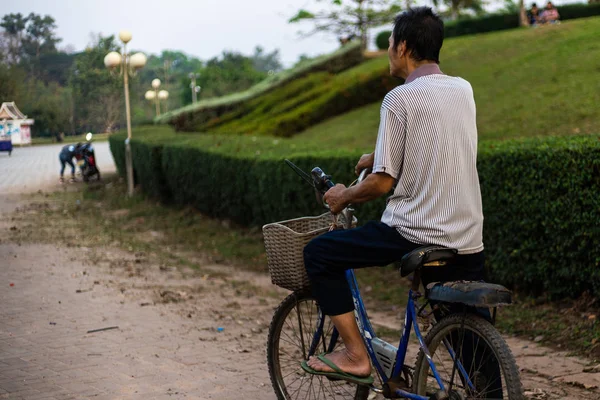 An old man rides an old bike through a park in Laos, Asia. back view