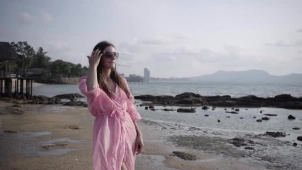 Mooie vrouw in een lange ontwikkeling van roze jurk wandelingen spinnen rondlopen op het strand op de rotsen. Close-up. 4k. — Stockvideo