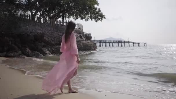 Mooie vrouw in een lange ontwikkeling van roze jurk wandelingen spinnen rondlopen op het strand op de rotsen. Close-up. 4k. — Stockvideo