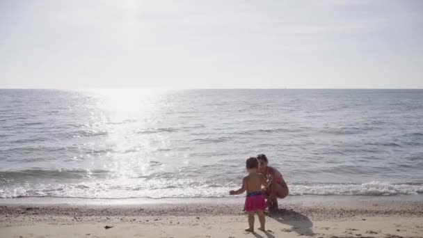 Madre e hija en trajes de baño rosados jugando en la arena a lo largo de la orilla del mar. concepto de una familia feliz. 4k . — Vídeos de Stock