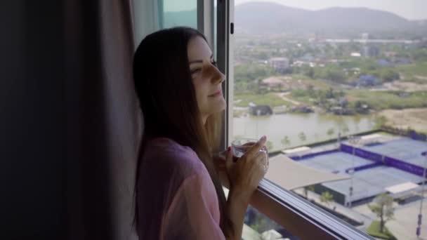 Femme en robe de chambre rose transparente se tient debout et regarde par la fenêtre panoramique et boit du café. 4k Hua Hin, Thaïlande . — Video