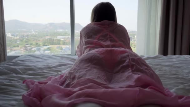 Femmina in una vestaglia trasparente rosa si trova sul letto e si affaccia sulla finestra panoramica e beve caffè. 4k Hua Hin, Thailandia — Video Stock